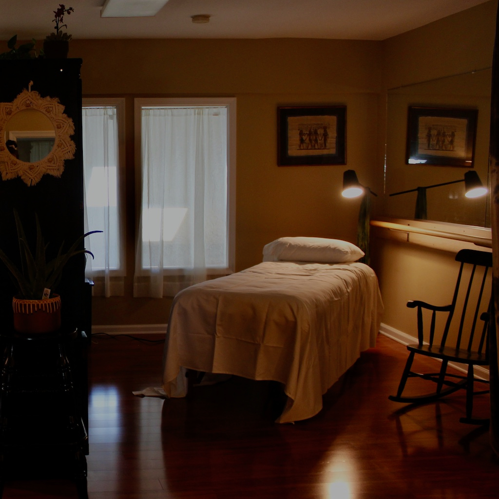 Picture of the Lwoer Bucks Acupuncture clinic. One acupuncture bed with lamplight shining on it, next to a rocking chair. Two windows overlooking the gym below with curtains covering. Black bookshelf on the left separating beds.