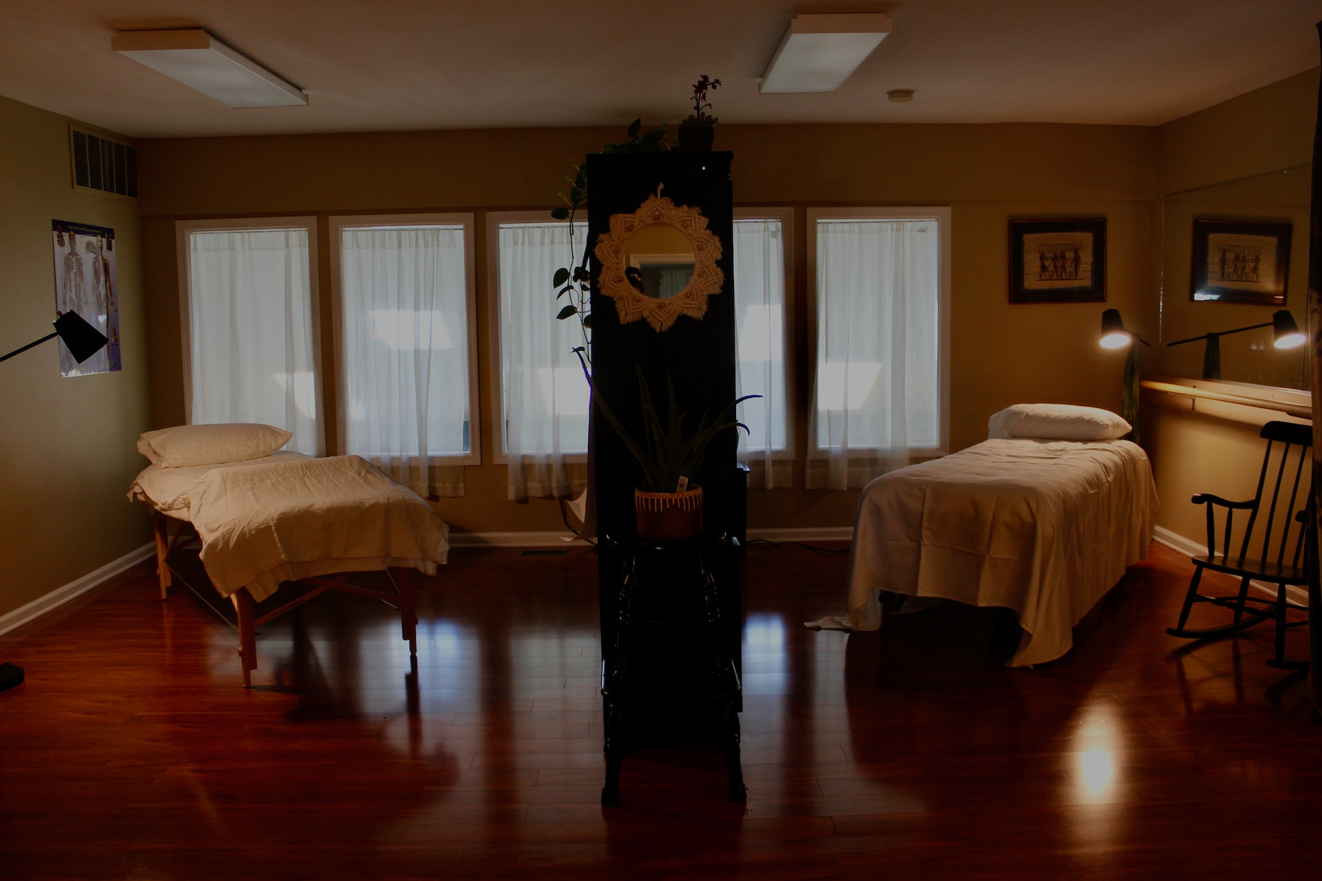 Picture of the Lower Bucks Acupuncture clinic. Two acupuncture beds separated by a large black bookshelf. Dark, shiny wooden floors. Lamplight shining on the beds. Five windows overlooking the gym on the lower floor with curtains.